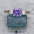 The "Dryope" ring in platinum, featuring a 1.13-carat Umba sapphire center stone, is displayed upright on a rough blue gemstone, highlighting the unique charm of this elegant three-stone design.