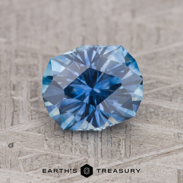 1.86-Carat Medium Blue Montana Sapphire (Heated) A 1.86-carat Medium Blue Montana Sapphire (Heated) is displayed faceted on a textured surface with the Earth’s Treasury logo beneath it.