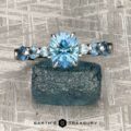 The "Minthe" ring in 14k white gold with a 1.39-carat Montana sapphire and smaller blue stones is displayed on a rough blue mineral, against a textured metallic background. The EARTH'S TREASURY logo appears at the bottom.