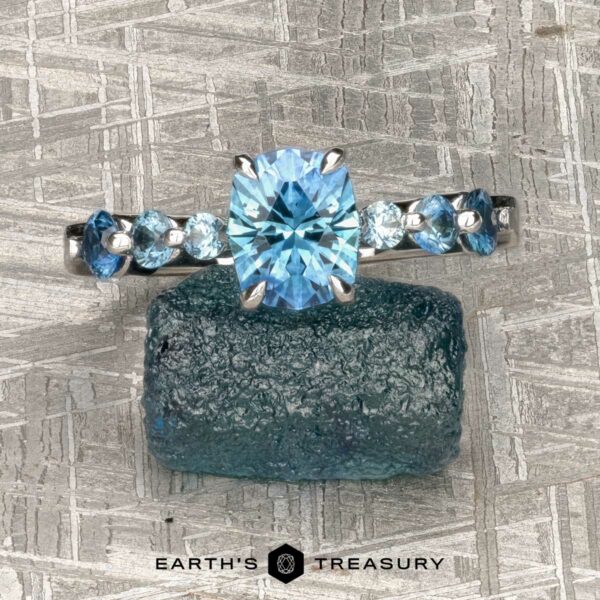 The "Minthe" ring in 14k white gold with 1.39-Carat Montana Sapphire The "Minthe" ring in 14k white gold with a 1.39-carat Montana sapphire and smaller blue stones is displayed on a rough blue mineral, against a textured metallic background. The EARTH'S TREASURY logo appears at the bottom.