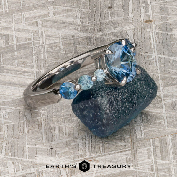 The "Minthe" ring in 14k white gold with 1.39-Carat Montana Sapphire The "Minthe" ring in 14k white gold with a 1.39-carat Montana sapphire is displayed on a rough blue mineral, set against a textured background featuring the Earth's Treasury logo at the bottom.