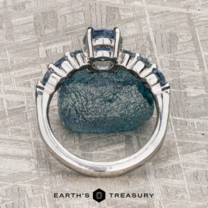 The "Minthe" ring in 14k white gold with a 1.39-carat Montana Sapphire is displayed upright against a rough blue stone on a textured gray surface. "EARTH'S TREASURY" is written below.