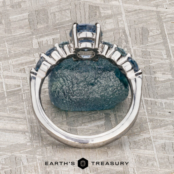 The "Minthe" ring in 14k white gold with 1.39-Carat Montana Sapphire The "Minthe" ring in 14k white gold with a 1.39-carat Montana Sapphire is displayed upright against a rough blue stone on a textured gray surface. "EARTH'S TREASURY" is written below.