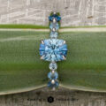 The "Minthe" ring in 14k white gold showcases a 1.39-carat Montana sapphire with round side stones, elegantly displayed on a green leaf. The Earth's Treasury logo is featured at the bottom.