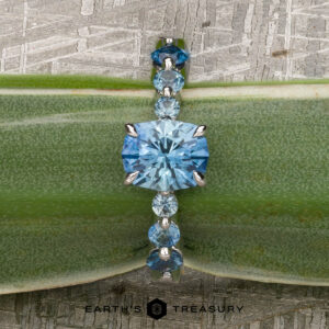 The "Minthe" ring in 14k white gold showcases a 1.39-carat Montana sapphire with round side stones, elegantly displayed on a green leaf. The Earth's Treasury logo is featured at the bottom.
