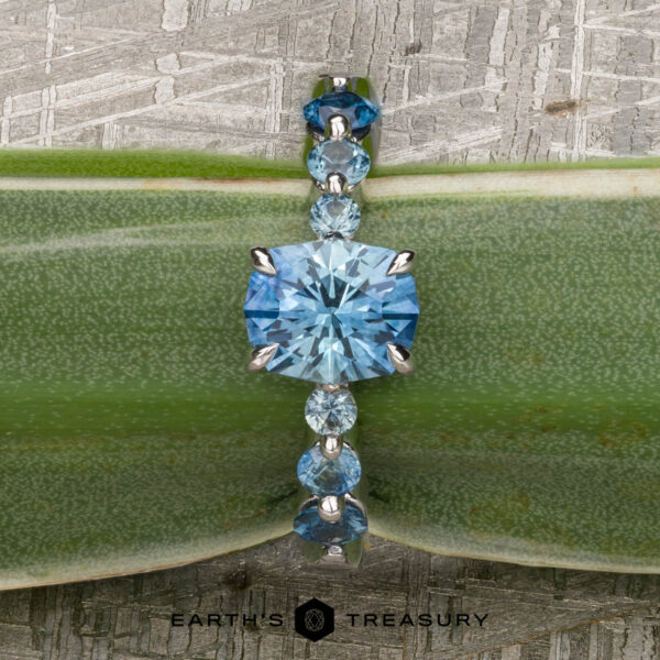 The "Minthe" ring in 14k white gold with 1.39-Carat Montana Sapphire The "Minthe" ring in 14k white gold showcases a 1.39-carat Montana sapphire with round side stones, elegantly displayed on a green leaf. The Earth's Treasury logo is featured at the bottom.