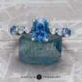 The "Minthe" ring in platinum, featuring a 1.60-carat Montana sapphire, stands upright on a rough blue crystal against a geometric background with the Earth’s Treasury logo below.