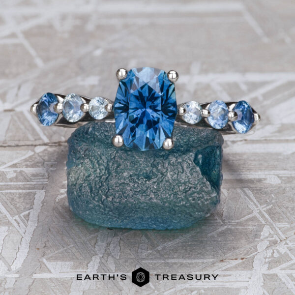 The "Minthe" ring in platinum with 1.60-carat Montana sapphire The "Minthe" ring in platinum, featuring a 1.60-carat Montana sapphire, stands upright on a rough blue crystal against a geometric background with the Earth’s Treasury logo below.