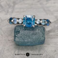The "Minthe" ring in 14k white gold showcases a 0.97-carat cushion-cut Montana sapphire centerpiece with round blue side stones, set atop a rough blue crystal and branded below with "Earth's Treasury.