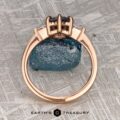 The "Melilai" Ring in 20k Pink Gold with a 1.42-carat Montana sapphire stands upright by a rough blue gemstone on textured gray, with the "EARTH'S TREASURY" logo shown below.