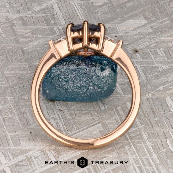 The "Melilai" Ring in 20k Pink Gold with 1.42-carat Montana sapphire The "Melilai" Ring in 20k Pink Gold with a 1.42-carat Montana sapphire stands upright by a rough blue gemstone on textured gray, with the "EARTH'S TREASURY" logo shown below.