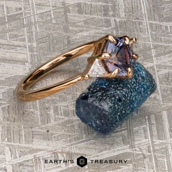 The "Melilai" Ring in 20k Pink Gold with 1.42-carat Montana sapphire The "Melilai" Ring in 20k pink gold, featuring a 1.42-carat Montana sapphire and two side stones, is displayed on a rough blue crystal atop a textured metallic surface.