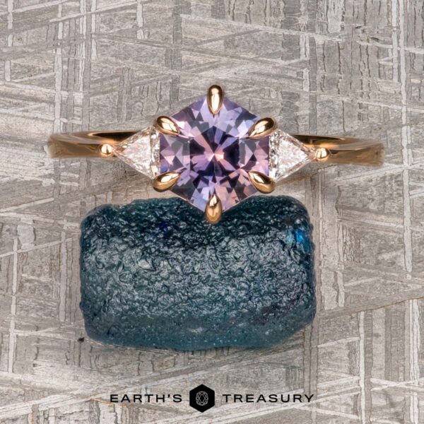 The "Melilai" Ring in 20k Pink Gold with 1.42-carat Montana sapphire The "Melilai" Ring in 20k pink gold with a 1.42-carat Montana sapphire is shown atop a rough blue gem, with two triangular side stones and "Earth's Treasury" text below on a textured background.