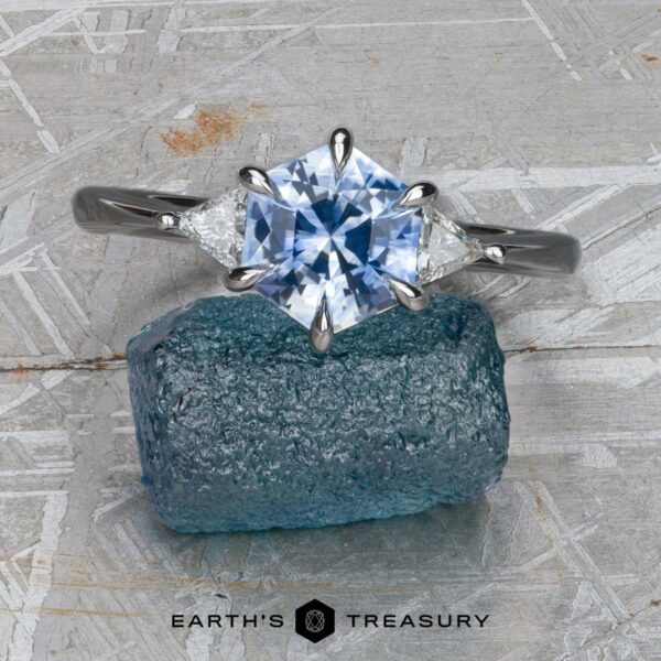 The "Melilai" ring in 18k white gold with 1.50-Carat Montana Sapphire The "Melilai" ring in 18k white gold, featuring a 1.50-carat Montana sapphire and two triangular diamonds, is displayed on rough blue mineral against a geometric gray background with the "Earth's Treasury" logo below.