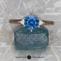 The "Melilai" ring in 14k rose gold, set with a 1.45-carat Montana sapphire and two side stones, is displayed on a rough blue mineral against a textured gray background.