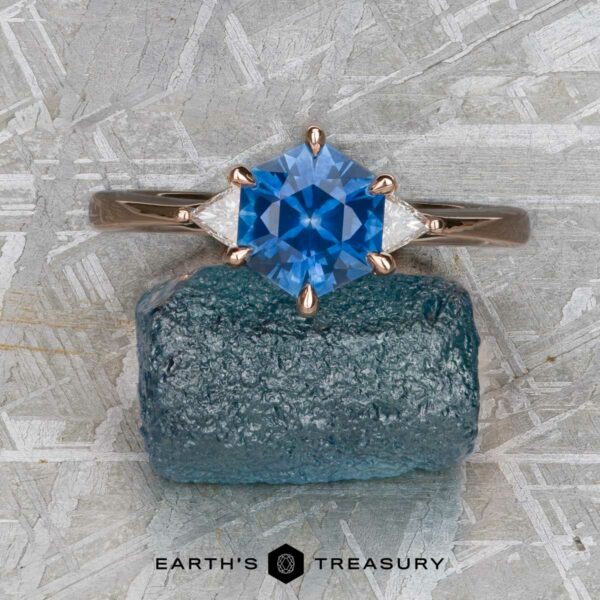 The "Melilai" in 14k rose gold with 1.45-Carat Montana Sapphire The "Melilai" ring in 14k rose gold, set with a 1.45-carat Montana sapphire and two side stones, is displayed on a rough blue mineral against a textured gray background.