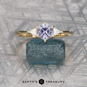 The "Melilai" ring in 14k yellow gold, featuring a 1.06-carat Montana sapphire and two clear side stones, is displayed on a rough blue-green mineral with the "EARTH'S TREASURY" logo below.