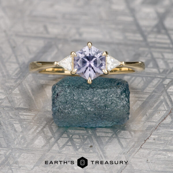 The "Melilai" ring in 14k yellow gold with 1.06-carat Montana sapphire The "Melilai" ring in 14k yellow gold, featuring a 1.06-carat Montana sapphire and two clear side stones, is displayed on a rough blue-green mineral with the "EARTH'S TREASURY" logo below.