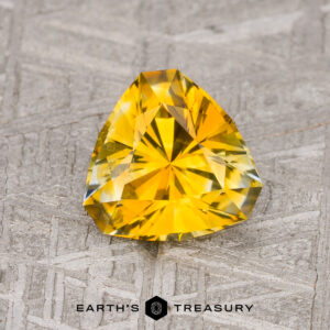 A 1.72-carat orange-yellow particolored Montana sapphire (heated), cut in a faceted triangle, displayed on a textured gray background with the "Earth's Treasury" logo at the bottom.
