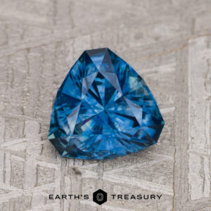 A 1.71-carat medium blue heated Montana Sapphire is shown on a textured surface, with the "Earth's Treasury" logo and text at the bottom.