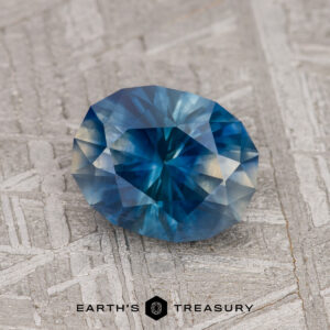 A 1.73-carat Blue-Teal-Cream Particolored Montana Sapphire (Heated) is showcased on a textured gray surface, with "EARTH'S TREASURY" displayed below.