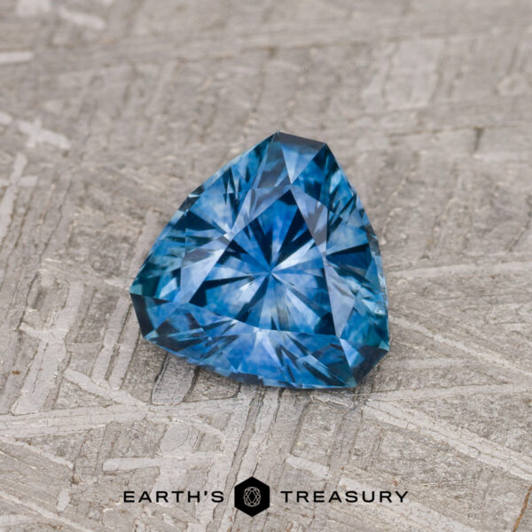 1.09-Carat Medium Blue Montana Sapphire (Heated) A 1.09-carat medium blue Montana Sapphire (heated), trillion-cut with intricate facets, rests on a textured gray surface above the text "EARTH'S TREASURY.