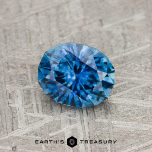 A 1.25-Carat Medium Blue Montana Sapphire (Heated) rests on a textured gray surface with "EARTH'S TREASURY" and a geometric logo beneath it.