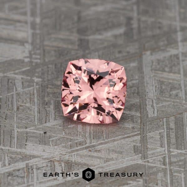 A 5.09-carat rich peach-pink Mahenge garnet, cut into a square cushion shape, is placed on a textured gray surface. The gemstone has multiple facets that beautifully reflect light. Below the gemstone, the text "Earth's Treasury" is visible.