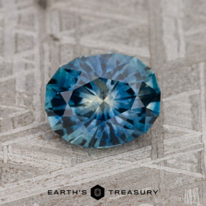 A 1.71-carat teal blue Montana Sapphire (heated) rests on a textured gray surface with the "EARTH'S TREASURY" logo below it.