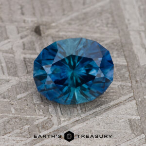 A 1.43-carat Peacock Blue Montana Sapphire (Heated), round and faceted, displays vivid blue-teal hues as it rests on a textured gray surface with the Earth’s Treasury logo beneath it.