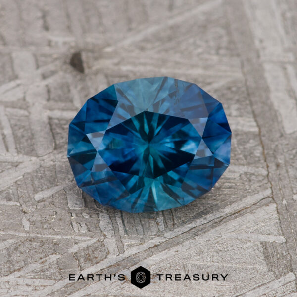 1.43-Carat Peacock Blue Montana Sapphire (Heated) A 1.43-carat Peacock Blue Montana Sapphire (Heated), round and faceted, displays vivid blue-teal hues as it rests on a textured gray surface with the Earth’s Treasury logo beneath it.