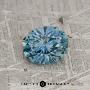 A 1.12-carat Robin's Egg Blue Montana Sapphire, round and faceted, rests on a textured gray surface. The "EARTH'S TREASURY" logo is displayed at the bottom of the image.