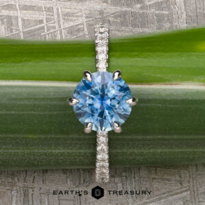 The Pave "Anemone" ring, featuring a 1.82-carat Montana sapphire set in 14k white gold, is displayed on a green stem. The "Earth's Treasury" logo appears at the bottom, showcasing this elegant piece.