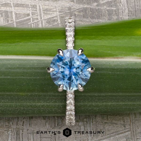 The Pave "Anemone" ring in 14k white gold with 1.82-carat Montana sapphire The Pave "Anemone" ring, featuring a 1.82-carat Montana sapphire set in 14k white gold, is displayed on a green stem. The "Earth's Treasury" logo appears at the bottom, showcasing this elegant piece.