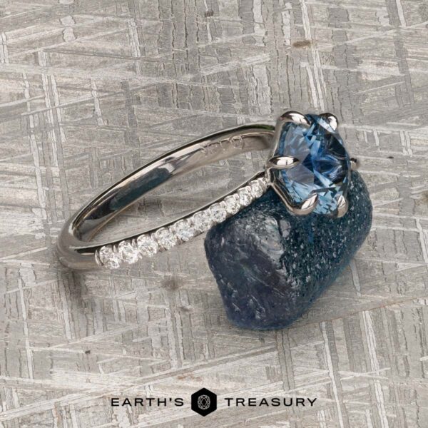 The Pave "Anemone" ring in 14k white gold with 1.82-carat Montana sapphire The Pave "Anemone" ring in 14k white gold, set with a 1.82-carat Montana sapphire, displayed as fine jewelry beside a rough blue crystal on textured gray; logo shows "Earth's Treasury.
