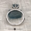 The Pave "Anemone" ring in 14k white gold with a 1.82-carat Montana sapphire is displayed upright on a textured blue stone, with "Earth's Treasury" at the bottom against a metallic, patterned background.
