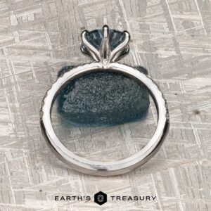 The Pave "Anemone" ring in 14k white gold with a 1.82-carat Montana sapphire is displayed upright on a textured blue stone, with "Earth's Treasury" at the bottom against a metallic, patterned background.