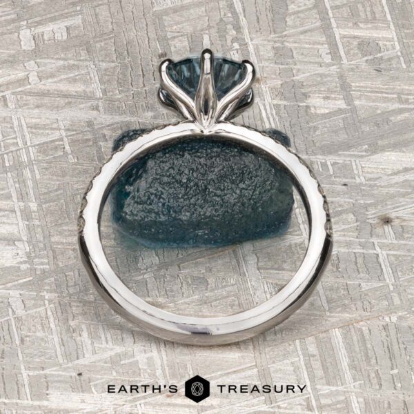 The Pave "Anemone" ring in 14k white gold with 1.82-carat Montana sapphire The Pave "Anemone" ring in 14k white gold with a 1.82-carat Montana sapphire is displayed upright on a textured blue stone, with "Earth's Treasury" at the bottom against a metallic, patterned background.