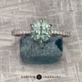 The Pave "Anemone" ring in platinum with a 1.66-carat Montana sapphire and diamond accents sits atop a rough green-blue stone on a textured gray surface, showcasing its designer elegance.