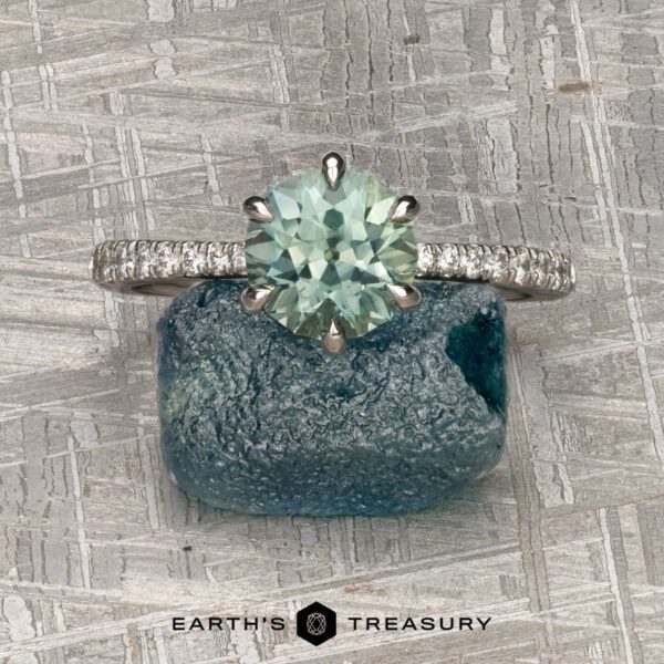 The Pave "Anemone" ring in platinum with 1.66-carat Montana sapphire The Pave "Anemone" ring in platinum with a 1.66-carat Montana sapphire and diamond accents sits atop a rough green-blue stone on a textured gray surface, showcasing its designer elegance.