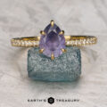 The Pave "Anemone" ring in 18K yellow gold, set with a 1.85-carat pear-shaped Umba sapphire, is displayed on a rough blue stone with the "Earth’s Treasury" logo beneath.