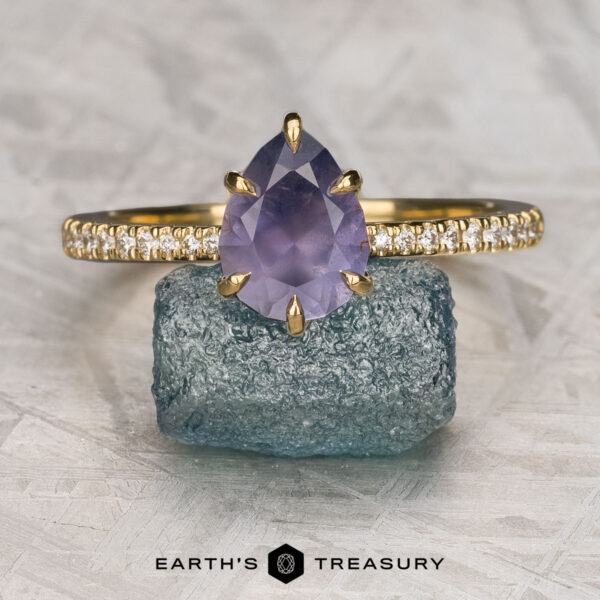 The Pave "Anemone" ring in 18K yellow gold with 1.85-carat Umba sapphire The Pave "Anemone" ring in 18K yellow gold, set with a 1.85-carat pear-shaped Umba sapphire, is displayed on a rough blue stone with the "Earth’s Treasury" logo beneath.