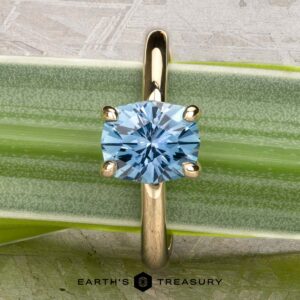 The "Alyssum" ring in 14k yellow gold, featuring a 1.63-carat Montana sapphire in a four-prong setting, rests on a green leaf—with the Earth’s Treasury logo below, inspired by alyssum blooms for an elegant touch.