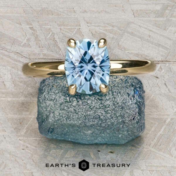 The "Alyssum" ring in 14k yellow gold with 1.63-Carat Montana Sapphire The "Alyssum" ring, crafted in 14k yellow gold with a 1.63-carat Montana sapphire, is displayed on a rough blue stone against a textured gray background.