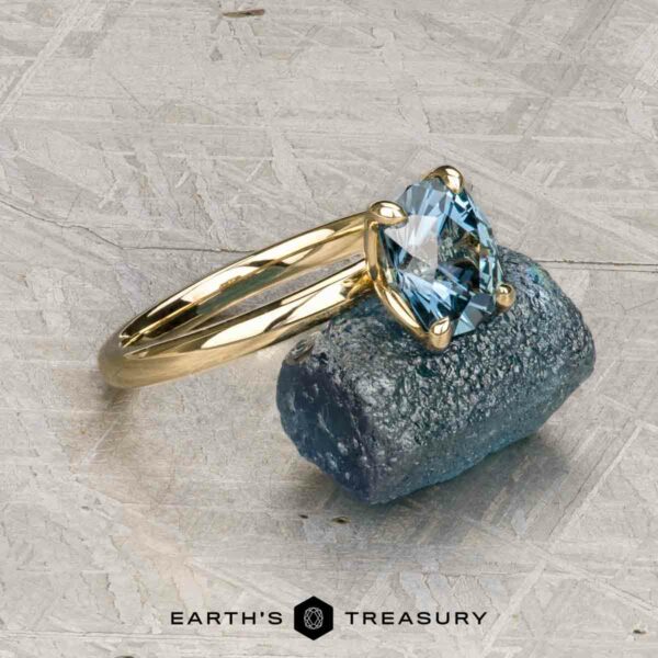 The "Alyssum" ring in 14k yellow gold with 1.63-Carat Montana Sapphire The "Alyssum" ring in 14k yellow gold with a 1.63-carat Montana sapphire rests on a rough blue crystal, with "Earth's Treasury" text below on a textured surface.