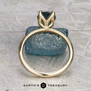 The "Alyssum" ring in 14k yellow gold with a 1.63-carat Montana Sapphire is displayed upright atop a rough blue stone, with the "EARTH'S TREASURY" logo featured below.
