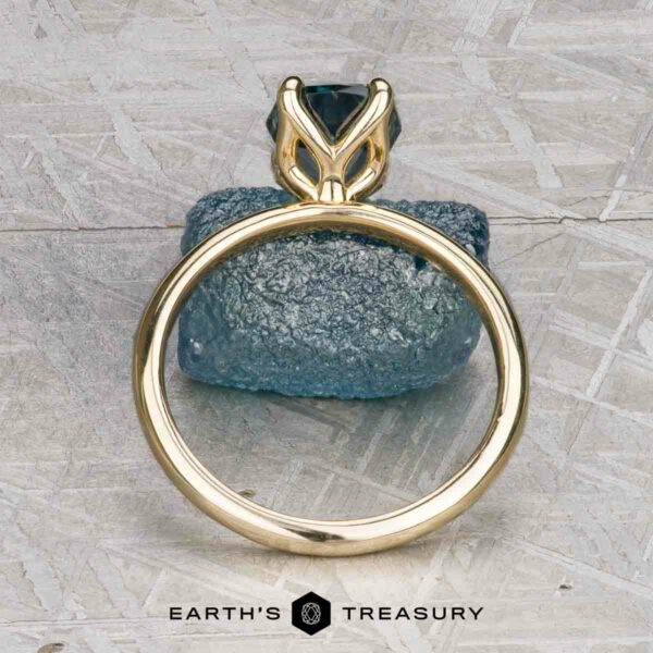 The "Alyssum" ring in 14k yellow gold with 1.63-Carat Montana Sapphire The "Alyssum" ring in 14k yellow gold with a 1.63-carat Montana Sapphire is displayed upright atop a rough blue stone, with the "EARTH'S TREASURY" logo featured below.