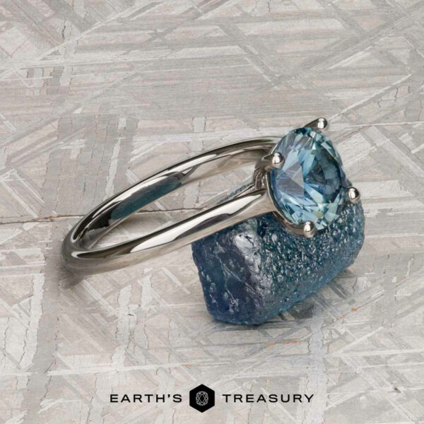 The "Cathedral" ring in platinum with 2.16-Carat Montana Sapphire A platinum "Cathedral" ring with a 2.16-carat Montana sapphire is set atop a rough blue stone on a patterned gray surface, highlighting its elegance. The Earthās Treasury logo appears at the bottom.
