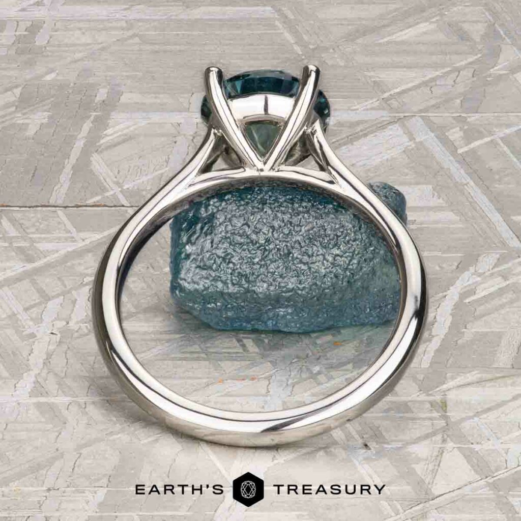 The "Cathedral" ring in platinum with a 2.16-carat Montana sapphire stands upright before a rough blue crystal on textured light gray, evoking solitaire card game elegance. "Earth's Treasury" is displayed at the bottom.