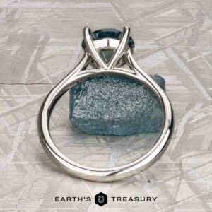 The "Cathedral" ring in platinum with a 2.16-carat Montana sapphire stands upright before a rough blue crystal on textured light gray, evoking solitaire card game elegance. "Earth's Treasury" is displayed at the bottom.